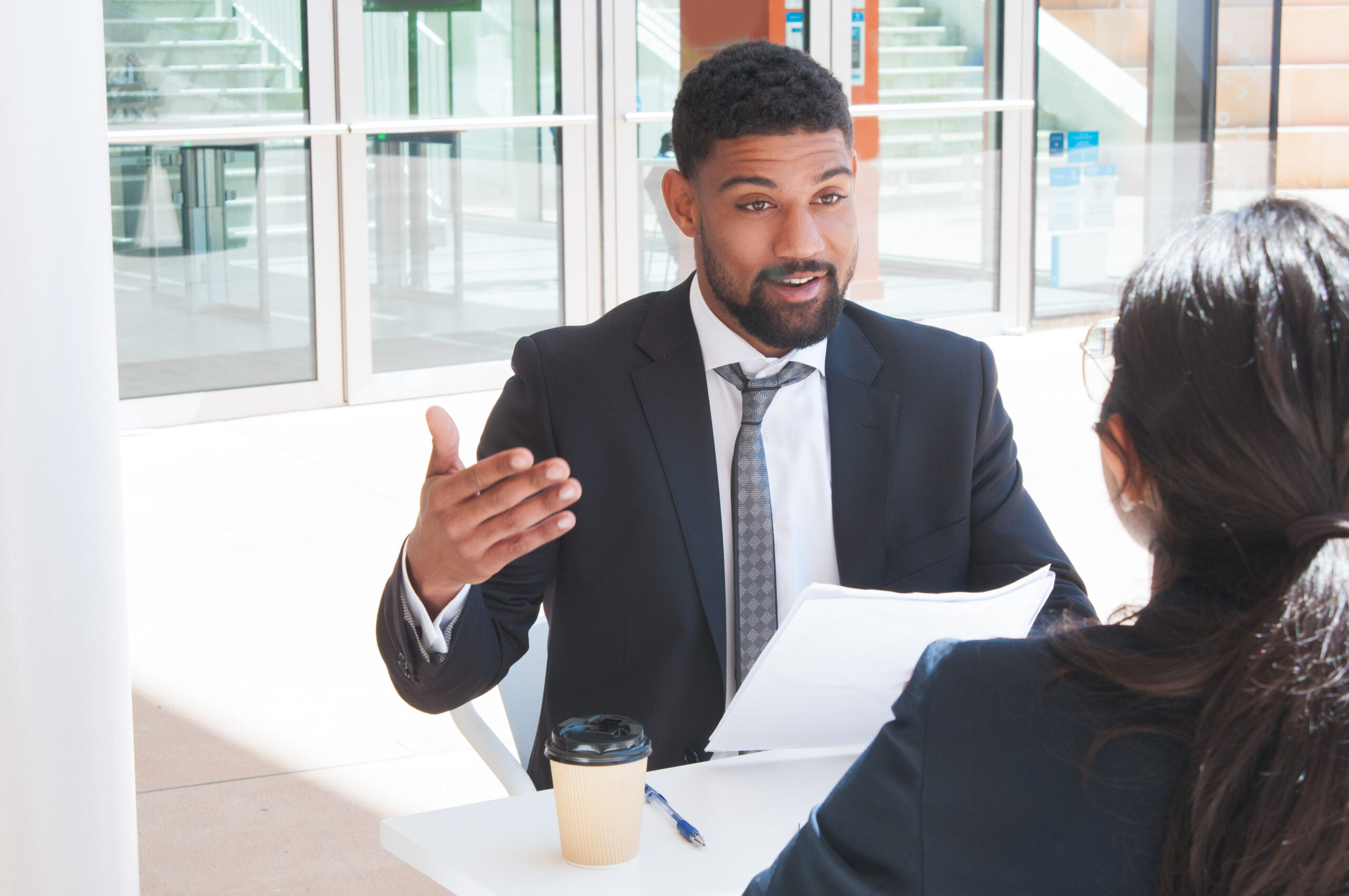 Businessman gesturing and talking with partner in outdoor cafe. Business man sitting and talking with woman who is sitting back to camera with building glass wall in background. Negotiations concept.